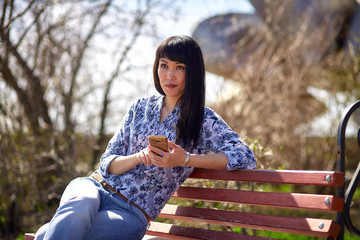 Beautiful Asian girl sitting on bench in park with phone in hand and looking into the distance