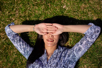 Beautiful Asian girl in a blue blouse lies on a green grass and covers her face with her hands