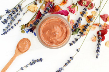 
Jar of facial bentonite clay mask amidst dry holistic herbs, top view white wooden table. Soft focus on skincare mud.