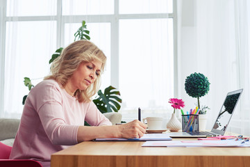 Cheerful madam of age 30-40 writing in notebook while sitting at table