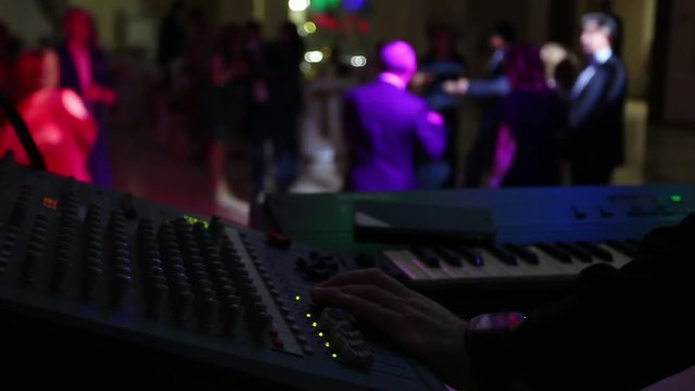 The musician in the foreground adjusts the sound and plays the keyboard instrument. People dance in the background.