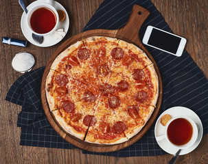 Pepperoni pizza standing on wooden table served with black textile napkin. Two cups of black tea and smartphone lying table surface. Flatlay