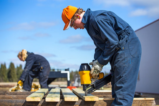 A teenage boy and a blurred teenage girl building outdoors with a power tool wearing protective industrial clothing