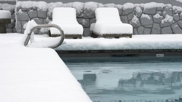 An outdoor swimming pool. the area around it and the lounging chairs covered in a thick layer, over half a foot, 20cm, of snow.