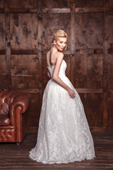Naklejka premium Fashion beauty bridal shooting. Beautiful fashion bride in wedding dress posing in front of woody background standing near brown leather sofa. studio shot in vogue style. 