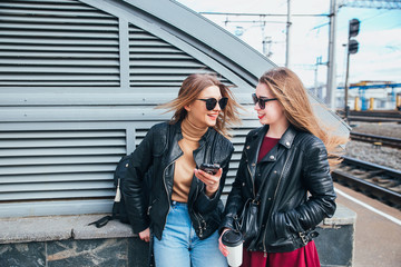 Fototapeta premium Two Women Talking in the City.Outdoor lifestyle portrait of two best friends hipster girls wearing stylish Leather Jacket and sunglasses with cofee, going crazy and having great time together