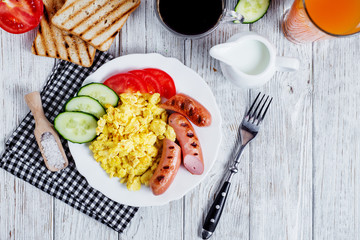 Hearty and traditional breakfast, scrambled eggs and milk with grilled pork, vegetables, tomato, cucumber, toast, juice and black coffee on a white wooden background