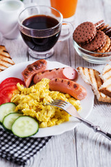 Hearty and traditional breakfast, scrambled eggs and milk with grilled pork, vegetables, tomato, cucumber, toast, juice and black coffee on a white wooden background