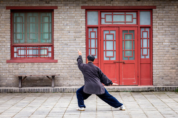 un homme chinois habillé en noir, réalise des figures de Kung fu, seul devant une maison chinoise