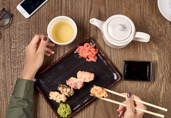 Flatlay. Closeup of woman hands with bamboo chopsticks eating sushi with green tea.