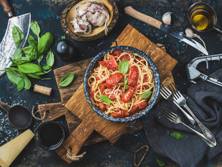 Italian style pasta dinner. Spaghetti with tomato and basil in plate on wooden board and ingredients for cooking pasta over dark plywood background, top view - obrazy, fototapety, plakaty