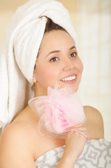Beautiful smiling young woman with white towel covering her head put a pink bath sponge under her cheek
