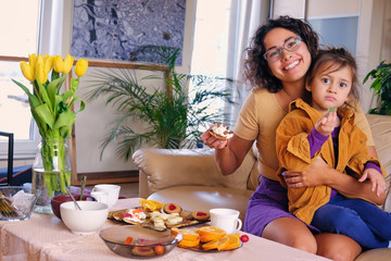 Obraz na płótnie Canvas Attractive brunette female in eyeglasses and her little cute daughter eats sweet cakes in a living room.