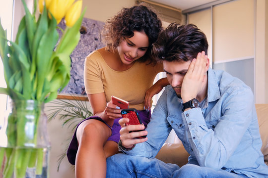 The attractive casual couple sits on a couch and using smartphones in a room.