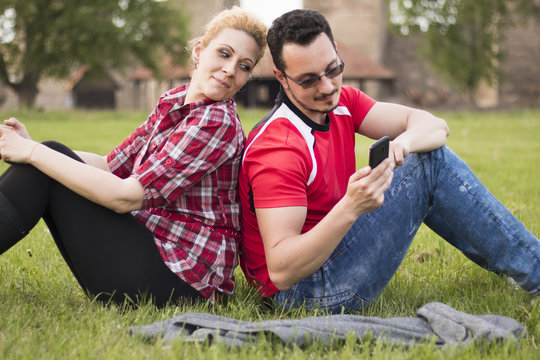 Girl and guy sitting back to back in grass, girl looking with smile over her shoulder at guy's telephone