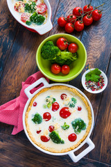 In process of broccoli and tomatoes quiche cooking, vegetables and cream-egg filling and half-baked pastry in the casseroles. Wooden rustic background, top view.