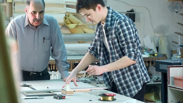 Young trainee trying to cut a glass for the frame, seniour worker watching
