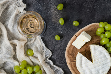 French soft cheese from Normandy region sliced on a wooden cut with green grapes and glass of white wine on dark rustic background, top view