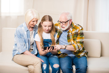 Cute girl with senior man and woman sitting on sofa and using smartphone