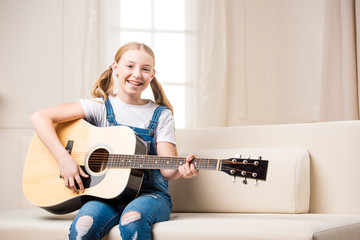 Cute smiling girl sitting on sofa and playing acoustic guitar