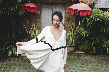 Young woman in white tunic in Ubud village with traditional balinese architecture. Style of Bali...