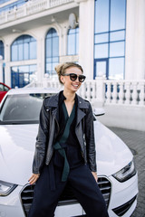 fashion outdoor photo of sexy beautiful woman with dark hair in black leather jacket and sunglasses posing in luxurious auto