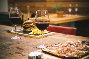 Traditional restaurant atmosphere with ham pizza and burger with chips in the background on rustic wooden table and glass of red wine