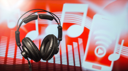 Large black headphones in front of a modern blurred background with notes, equalizer and a smartphone icon. An abstract online music streaming and listening concept with free copy space for text. 