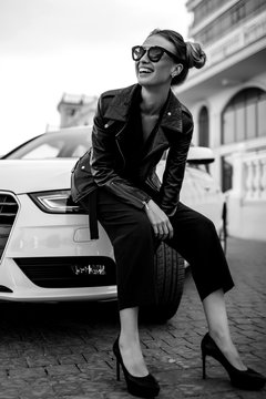 fashion outdoor photo of sexy beautiful woman with dark hair in black leather jacket and sunglasses posing in luxurious auto