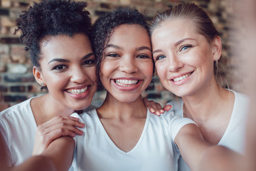 Beautiful and funny girls in white T-shirts makes selfie. Two afro-americans and one european.