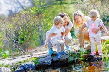 Two mothers laugh while holding their children by a small pond for a walk in the park