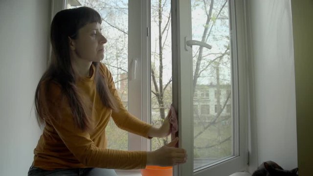 Young Brunette Woman In A Yellow Sweater Washes A Window In Her Apartment Using A Pink Rag On A Sunny Spring Day.