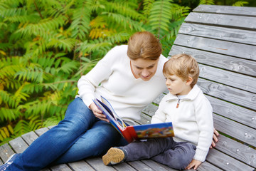 Young mother and toddler boy reading book in autumn park.