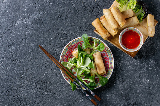 Fried spring rolls with red pepper sauces, served in traditional china plate with fresh green salad and wooden chopsticks over black texture background. Flat lay, space. Asian food