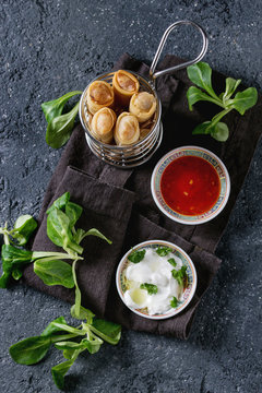 Fried spring rolls with red and white sauces, served in traditional china plate and fries basket with fresh green salad over black texture background. Flat lay, space. Asian food