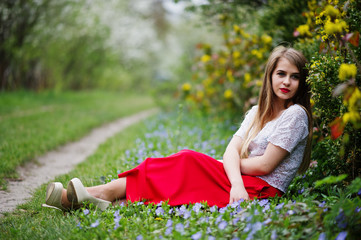 Portrait of sitiing beautiful girl with red lips at spring blossom garden on grass with flowers, wear on red dress and white blouse.