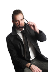 Attractive man sitting down in front of a white background wearing a shirt, tie, jeans and a black coat, holding a pair of sunglasses looking at camera.