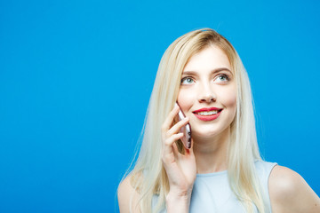 Fototapeta premium Portrait of Smiling Blonde with Sensual Lips Talking with Somebody by Her Smartphone on Blue Background. Cute Girl with Mobile in Studio.