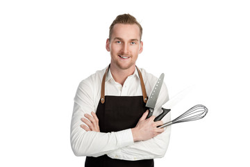 Handsome smiling chef in white and black, holding 2 knifes and a whisk looking at camera standing against a white background.