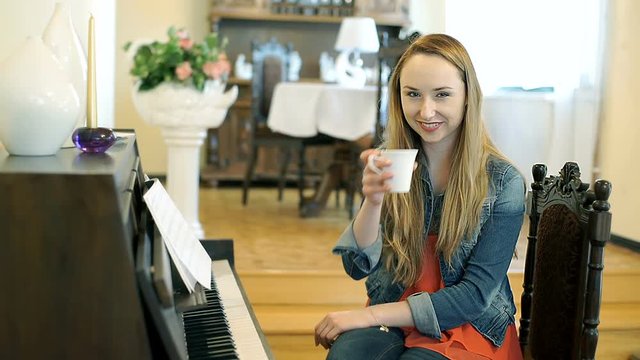 Pretty girl drinking tea and smiling to the camera while sitting next to the piano
