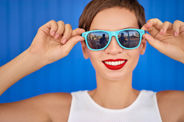 Colorful portrait of  happy young brunette woman with bright makeup holding sunglasses. Blue background.