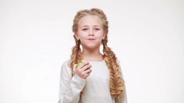 Young adorable Caucasian girl with long pretty blonde hair eating apple and smiling on white background in slowmotion