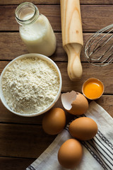 Baking ingredients flour, cracked eggs, open yolk, milk, rolling pin, linen towel, rustic kitchen interior, utensils, top view