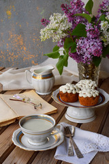 Romantic composition of  bouquet white and purple lilacs, cupcakes with curd cream, cups tea with milk, old book on wooden background.