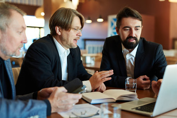 Three concentrated businessmen in suits sitting around cafe table and discussing statistical data, waist-up portrait