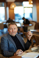 Waist-up portrait of mature manager with slight smile distracted from work in order to pose for photography, spacious cafe with guests on background