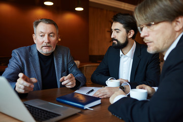 Waist-up portrait of bearded entrepreneurs analyzing risks and benefits of project investment with help of modern laptop while gathered together in dim cafe