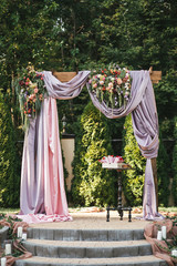 Arch for the wedding ceremony in the summer on the street, decorated with fresh flowers, eucalyptus, candles and a pink cloth. Against the background of trees and green plants