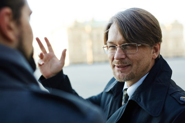 Bearded middle-aged manager looking at his colleague with squinty eyes while talking to him outdoors, over shoulder view