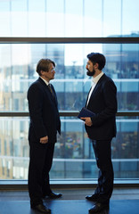 Full-length portrait of confident colleagues in suits gathered together against panoramic window of office lobby and analyzing results of completed work, profile view